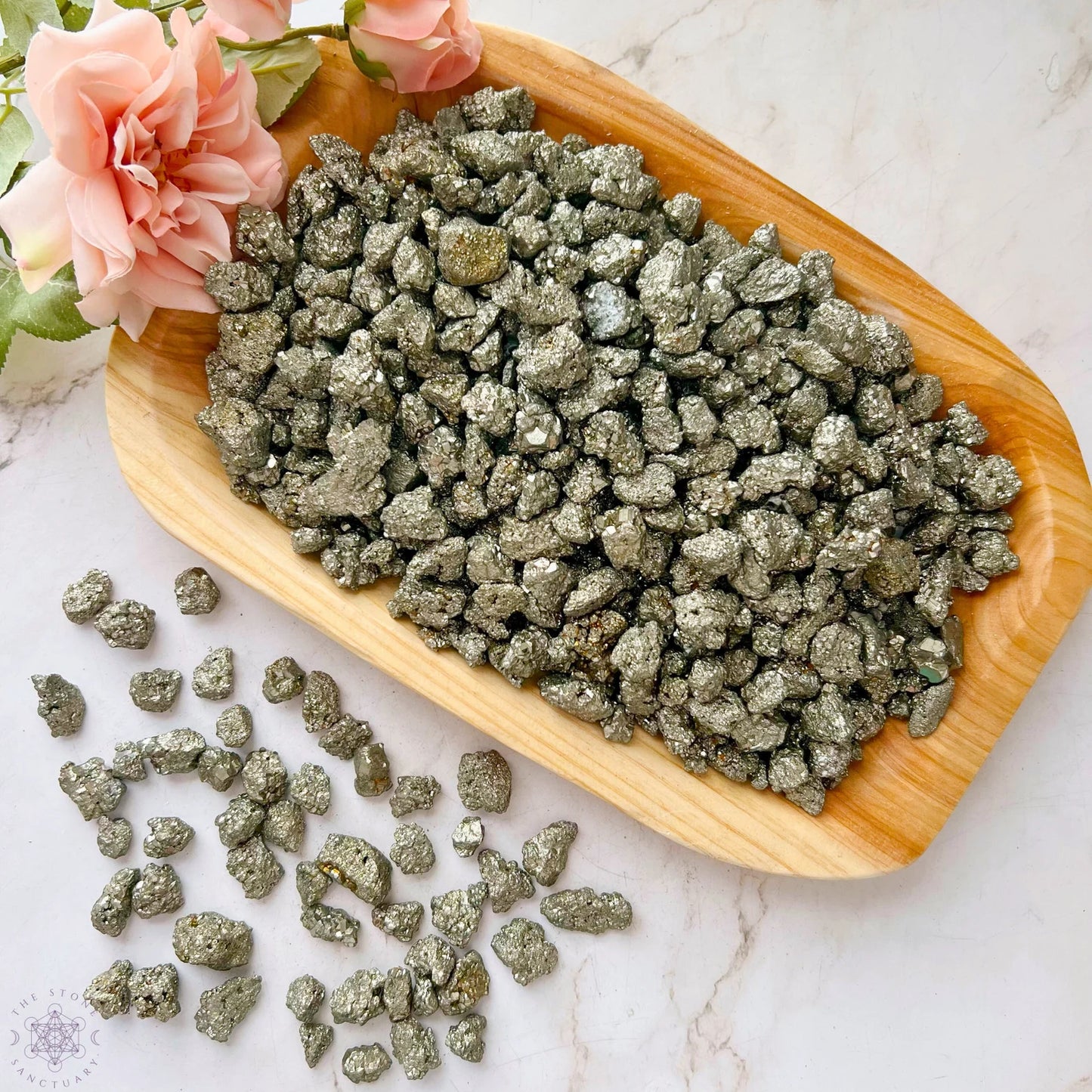 A wooden tray filled with shiny, metallic 12-18mm Pyrite Crystal Chips is placed on a marble surface. Some raw and unpolished stones are scattered beside the tray. Pink roses are positioned at the top left corner, partially in the frame.