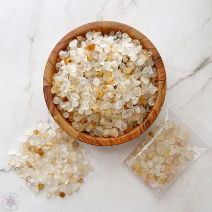 A wooden bowl filled with small, translucent Golden Healer Quartz Crystal Chips rests on a marble surface. Some crystal chips are scattered beside the bowl, while a plastic bag containing more Golden Healer Quartz Crystal Chips lies nearby, infusing the scene with the serene energy of Universal Life Force.