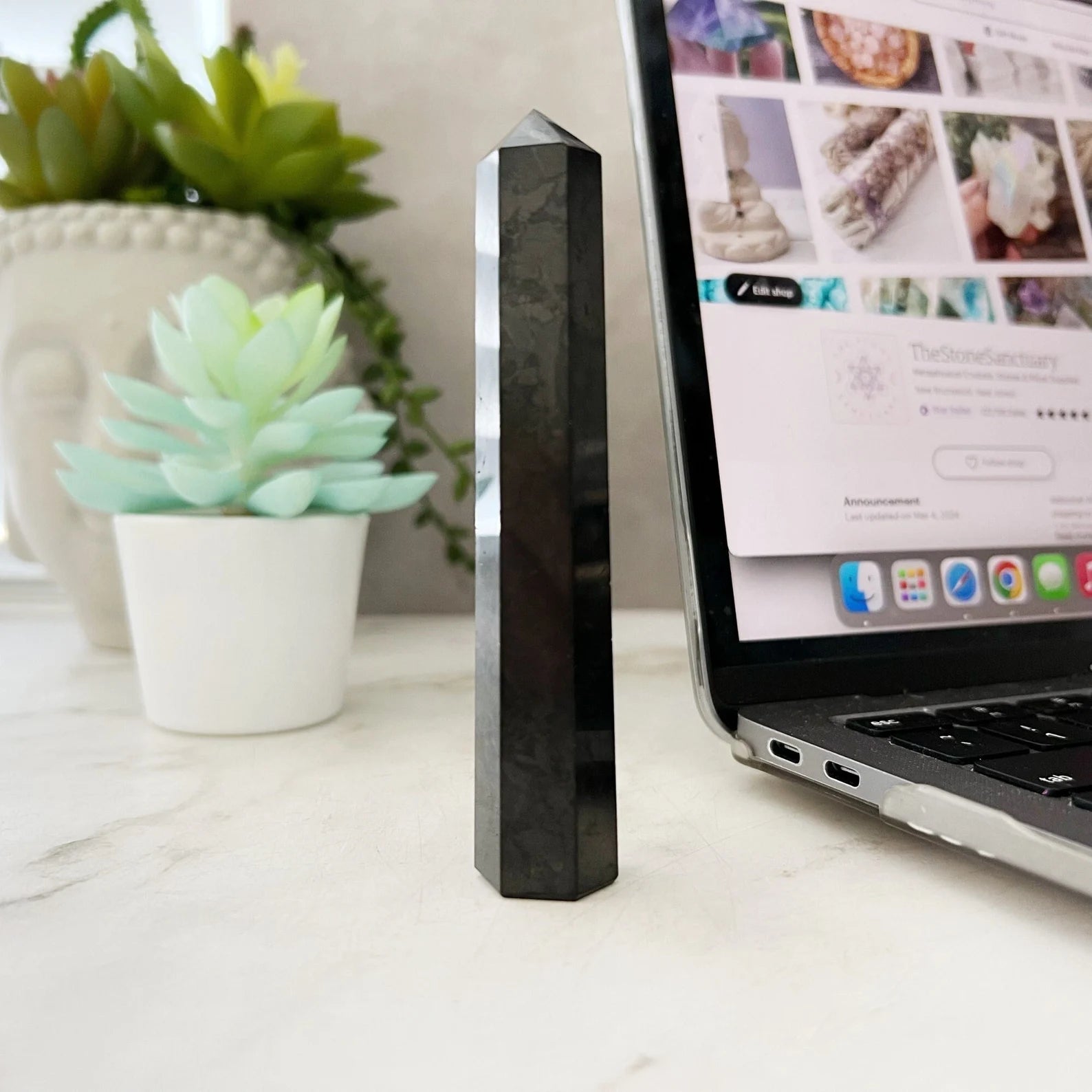 A sleek, hexagonal 3-4" Shungite Point stands upright on a white surface next to a succulent plant in a white pot. Part of an open laptop displaying web pages about crystal energy work and electromagnetic frequencies is visible in the background.