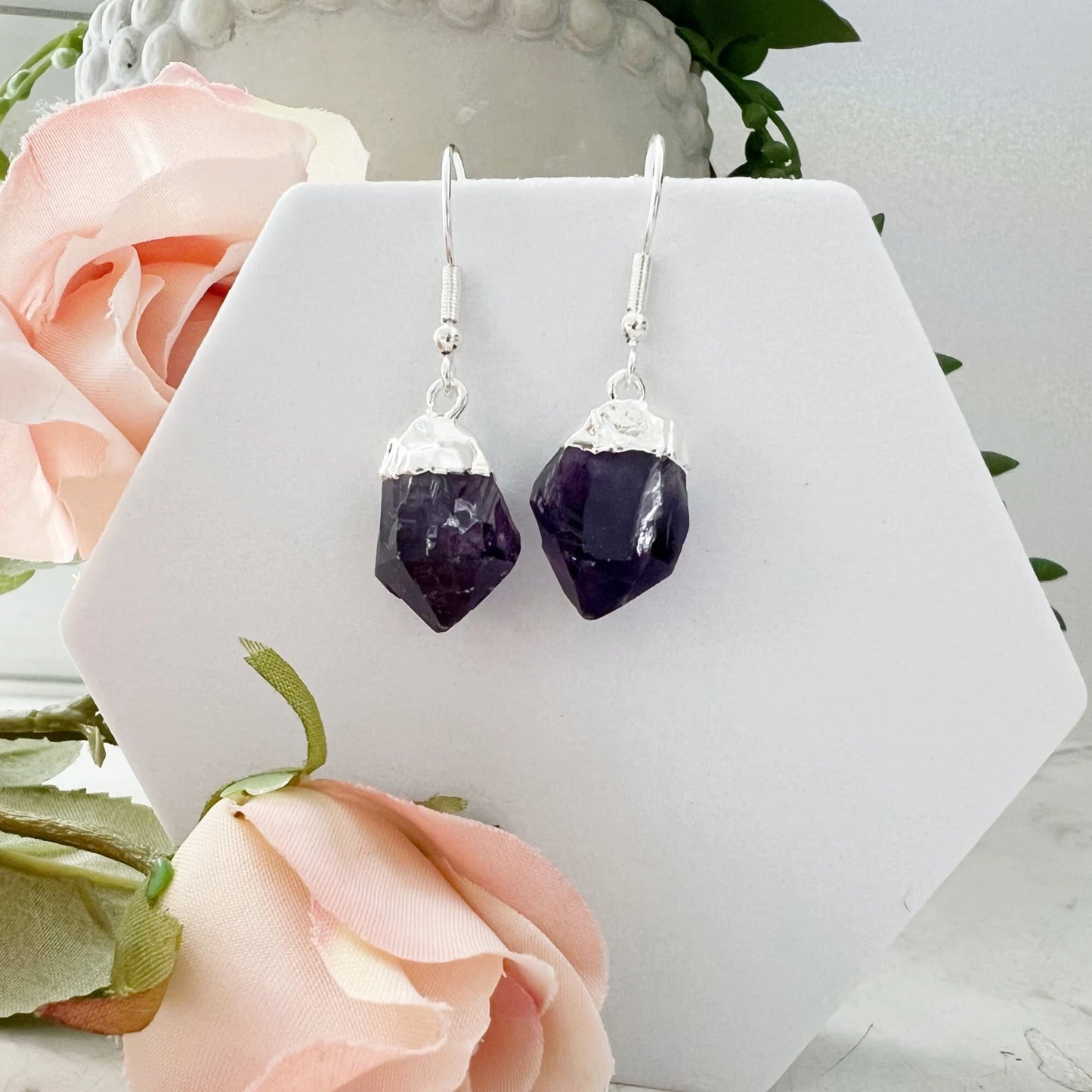 A pair of Raw Amethyst Point Dangle Earrings with silver hooks is displayed on a white hexagonal stand. Soft pink roses are placed around the base of the stand, with a decorative textured vase in the background. The deep purple amethyst provides an elegant and serene touch to the setting.