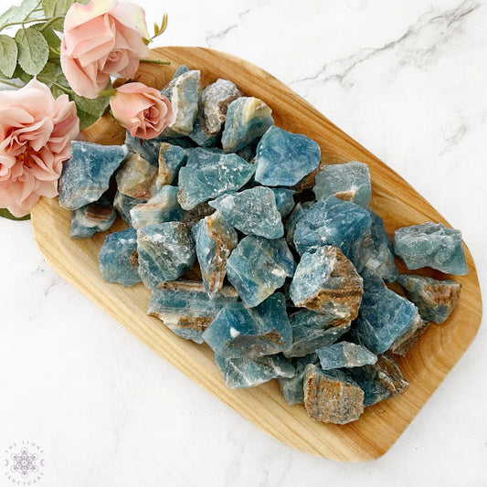 A wooden tray holds several large, unpolished Raw Blue Onyx Stones. Pink roses rest in the upper left corner of the tray, adding a contrasting touch of color. The tray is placed on a white marble surface.