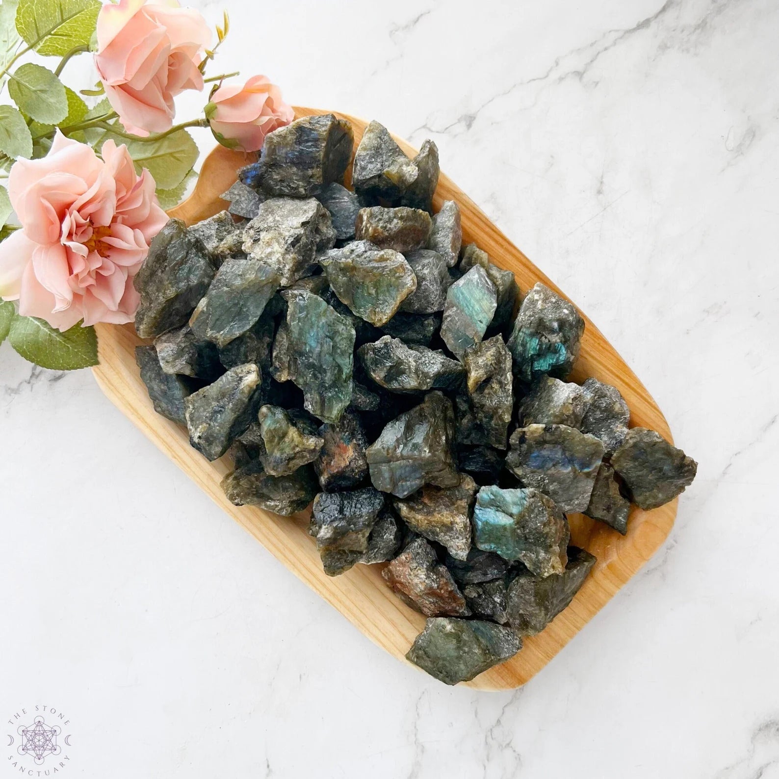 A wooden tray filled with Raw Labradorite Stones, showcasing their labradorescence in iridescent hues of blue, green, and gold, is placed on a white marble surface. Pink roses with green leaves lie next to the upper left side of the tray.