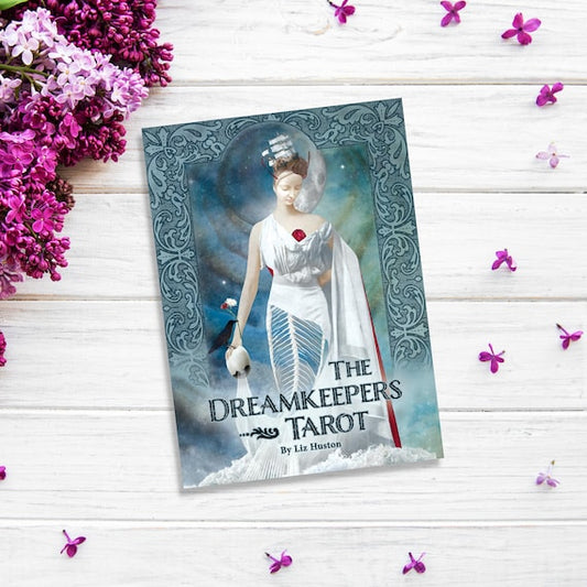 An image of "The Dreamkeepers Tarot" by Liz Huston, placed on a white wooden surface surrounded by purple flowers. The tarot deck cover features an ethereal figure in white robes holding a blue mask, set against a mystical background with ornate designs from The Dreamkeepers Tarot.