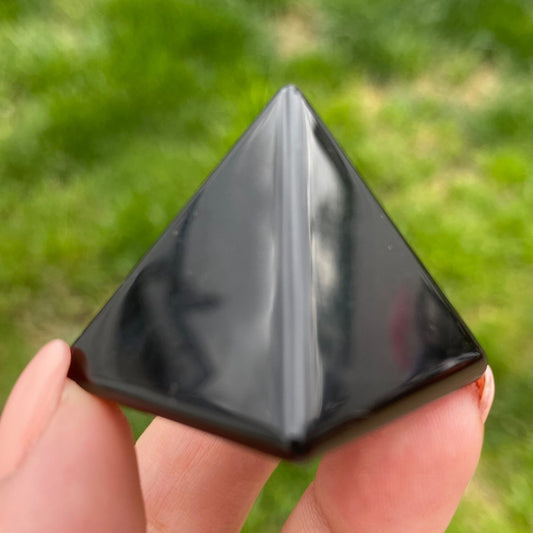 Close-up of a hand holding a 1" Mini Obsidian Pyramid against a background of green grass. The pyramid has an even, glossy surface that reflects light. This protective talisman gleams under natural lighting in the outdoor scene.