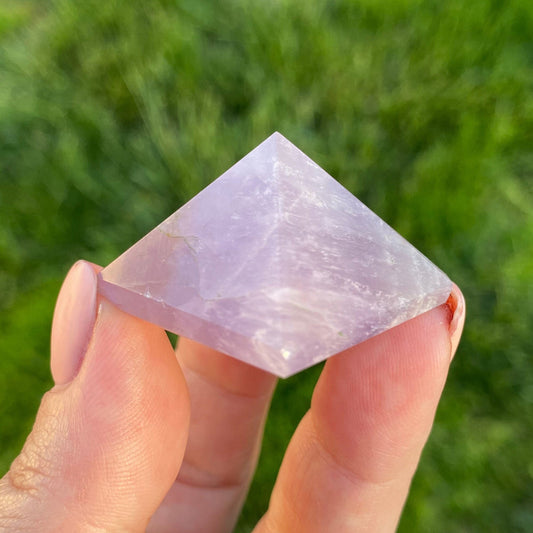 A close-up of a hand holding a small, purple 1" Mini Amethyst Pyramid against a blurred green grassy background. The crystal has a translucent quality with visible internal patterns and textures, evoking thoughts of its profound metaphysical properties and spiritual journey.