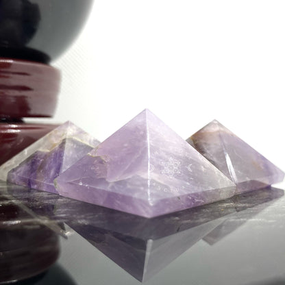 A close-up of a hand holding a small, purple 1" Mini Amethyst Pyramid against a blurred green grassy background. The crystal has a translucent quality with visible internal patterns and textures, evoking thoughts of its profound metaphysical properties and spiritual journey.