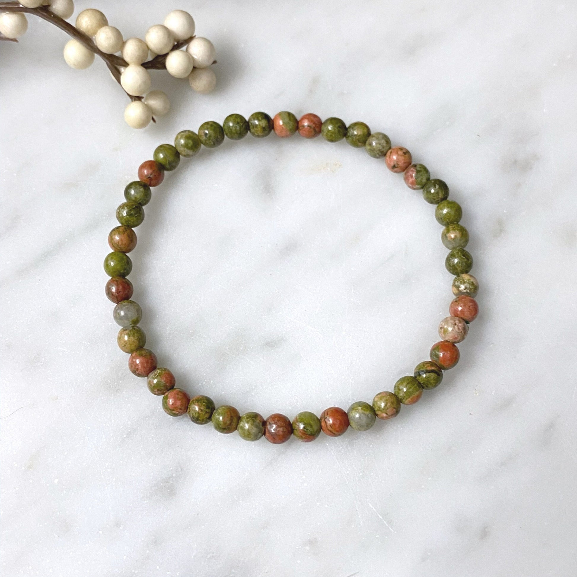 A 4mm Unakite Beaded Bracelet made of round, multicolored stones in shades of green and pink is displayed against a glossy black background, creating a reflection underneath.