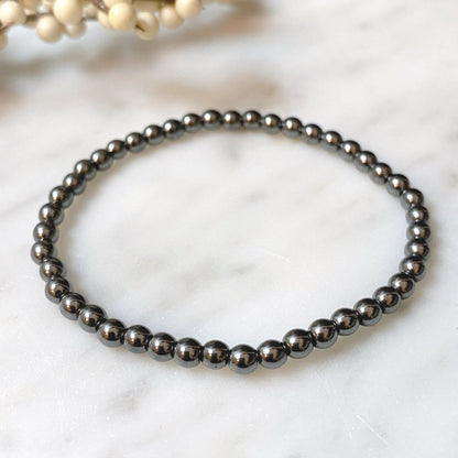 A close-up of a 4mm Hematite Beaded Bracelet made of shiny, reflective black beads. In the blurred background, there are small white spherical decorative elements. The overall tone of the image is elegant and minimalist, highlighting the grounding properties of the polished hematite beads.