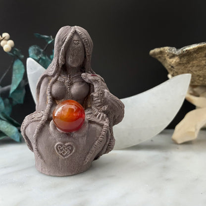 A Tiamat Goddess Statue of a faceless woman with long hair, seated cross-legged, holding a red gemstone orb. The figurine, made from natural gypsum, has a Celtic knot etched on its base and is accompanied by a white crescent shape and other decorative elements in the background.