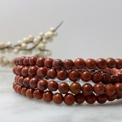 A close-up of a multi-layered 4mm Red Jasper Beaded Bracelet made of polished beads, arranged in a spiral. Behind the bracelet, a blurred branch adorned with small white berries enhances the minimalistic and natural aesthetic, highlighting the spiritual properties associated with Red Jasper.