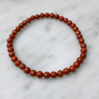 A close-up of a multi-layered 4mm Red Jasper Beaded Bracelet made of polished beads, arranged in a spiral. Behind the bracelet, a blurred branch adorned with small white berries enhances the minimalistic and natural aesthetic, highlighting the spiritual properties associated with Red Jasper.