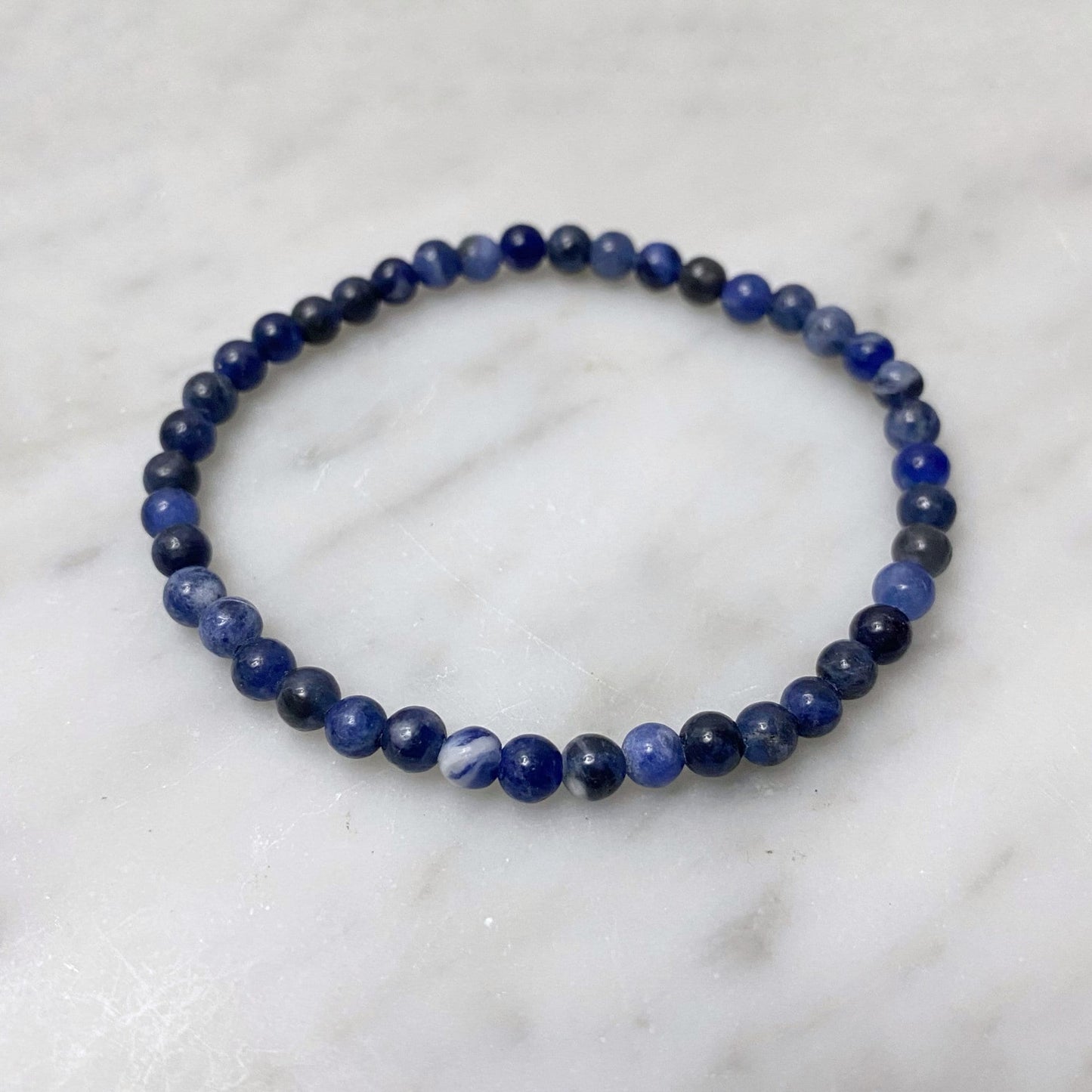 A stack of 4mm Sodalite Beaded Bracelets is displayed on a white marble surface. The deep blue beads have slight variations in color, adding texture to the bands. In the blurred background, delicate white buds on branches add a soft contrast, promoting emotional balance and healing Usui Reiki energy.