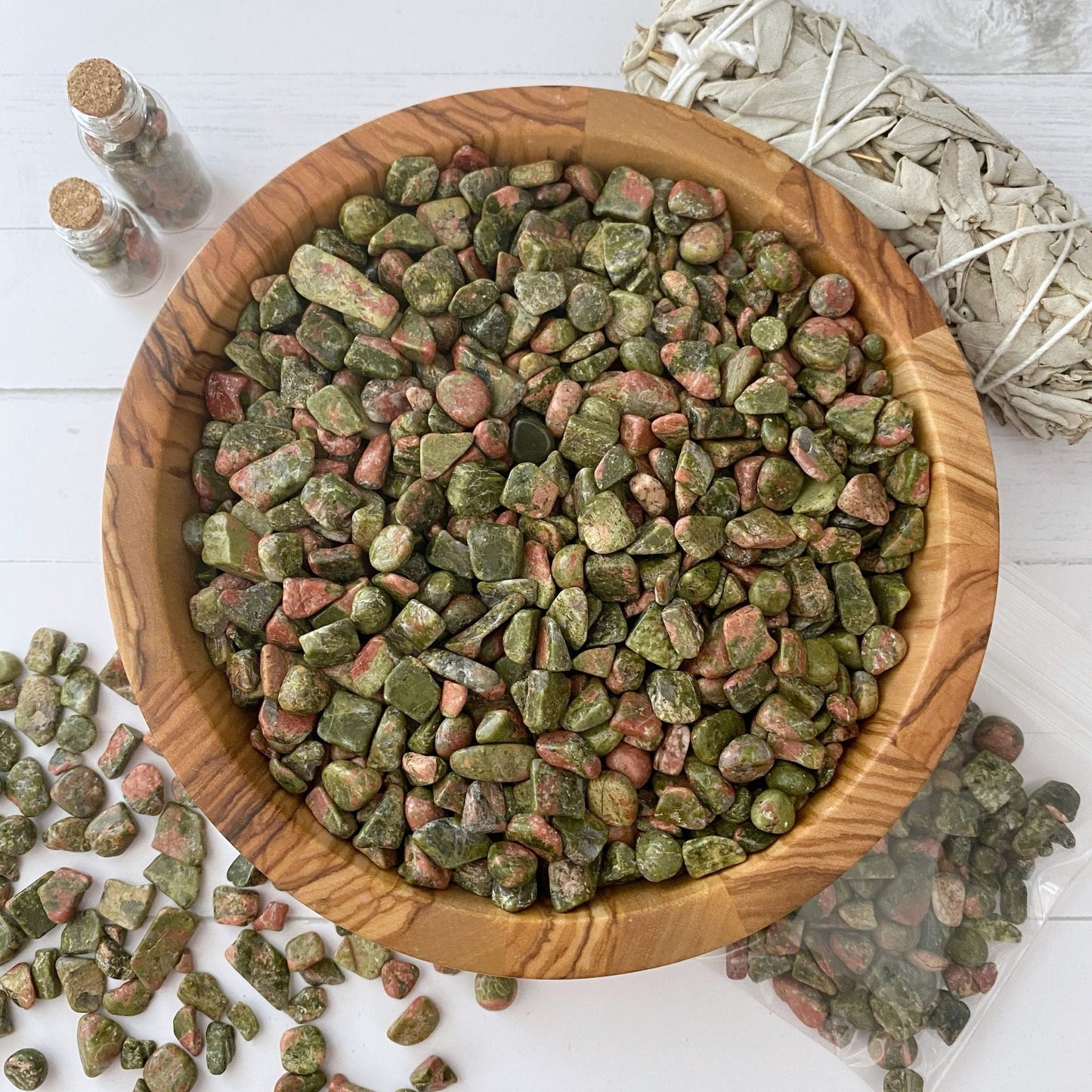 A wooden bowl filled with green and pink Unakite Crystal Chips is placed on a white surface. Some Unakite Crystal Chips are scattered outside the bowl. Small, corked glass bottles and a tied bundle of white sage, perfect for candle-making projects, are also present in the background.