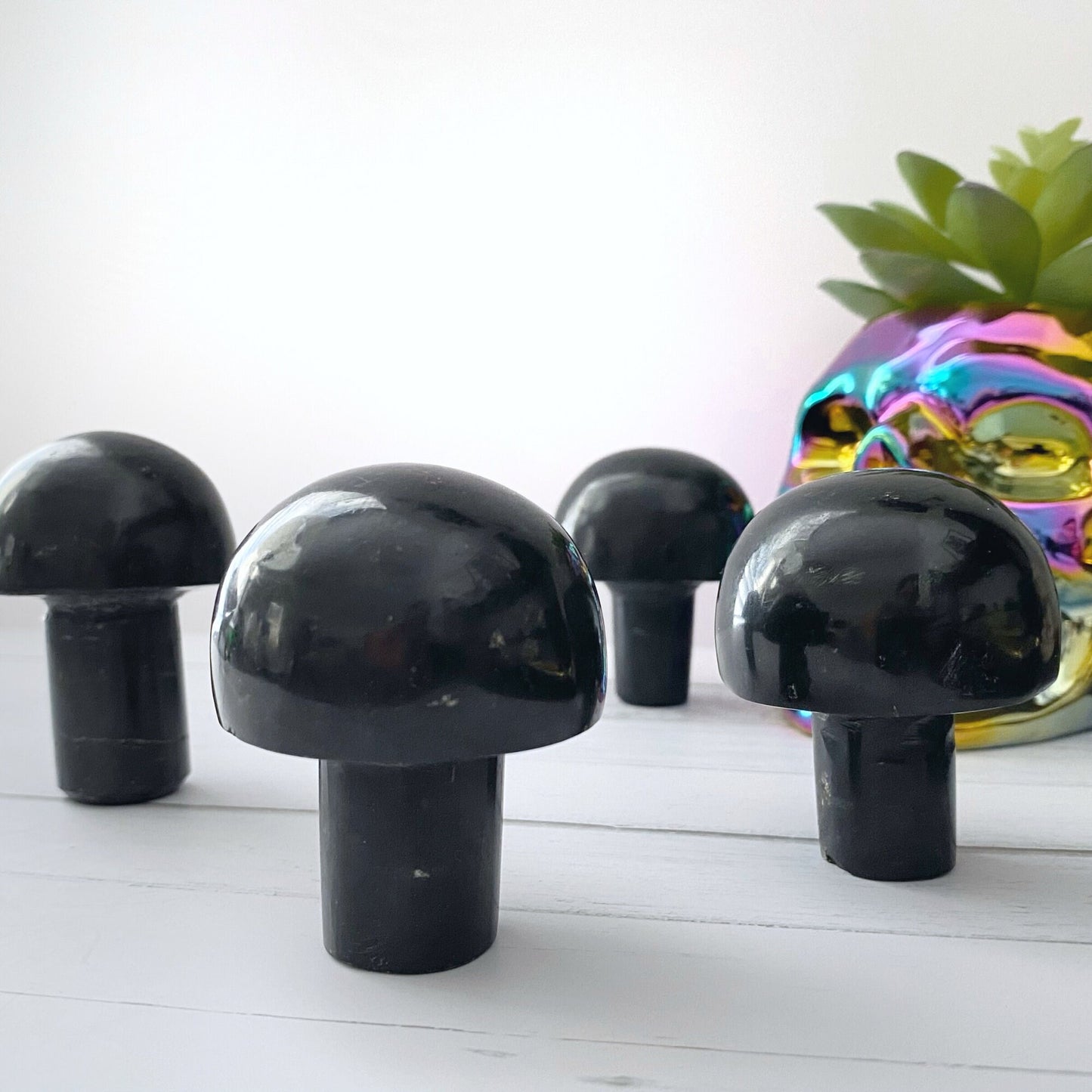 Four 2" Black Tourmaline Mushroom Massagers are placed on a white wooden surface, with an iridescent skull-shaped container in the background, partially obscured by a green succulent plant.