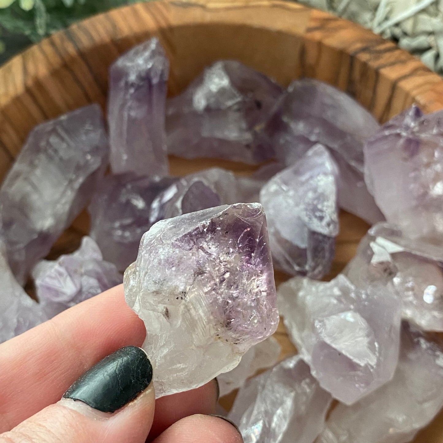 A wooden bowl filled with 1-2" Raw Amethyst Points is placed on a wooden surface. To the right of the bowl is a bundle of sage, and to the left, part of a green leafy plant is visible. The overall setting has a natural and earthy feel, enhanced by the metaphysical properties of the amethyst.