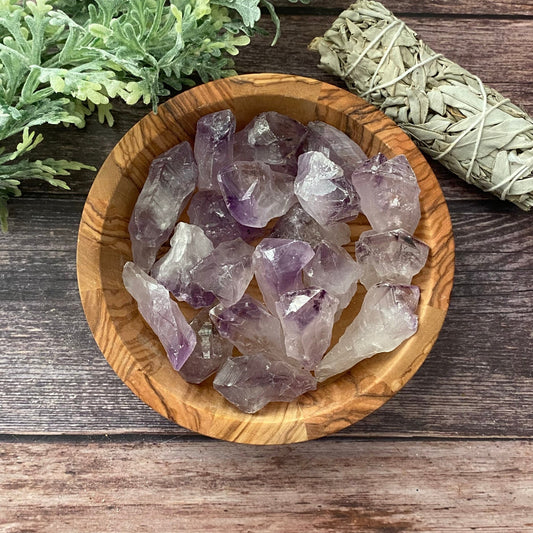 A wooden bowl filled with 1-2" Raw Amethyst Points is placed on a wooden surface. To the right of the bowl is a bundle of sage, and to the left, part of a green leafy plant is visible. The overall setting has a natural and earthy feel, enhanced by the metaphysical properties of the amethyst.