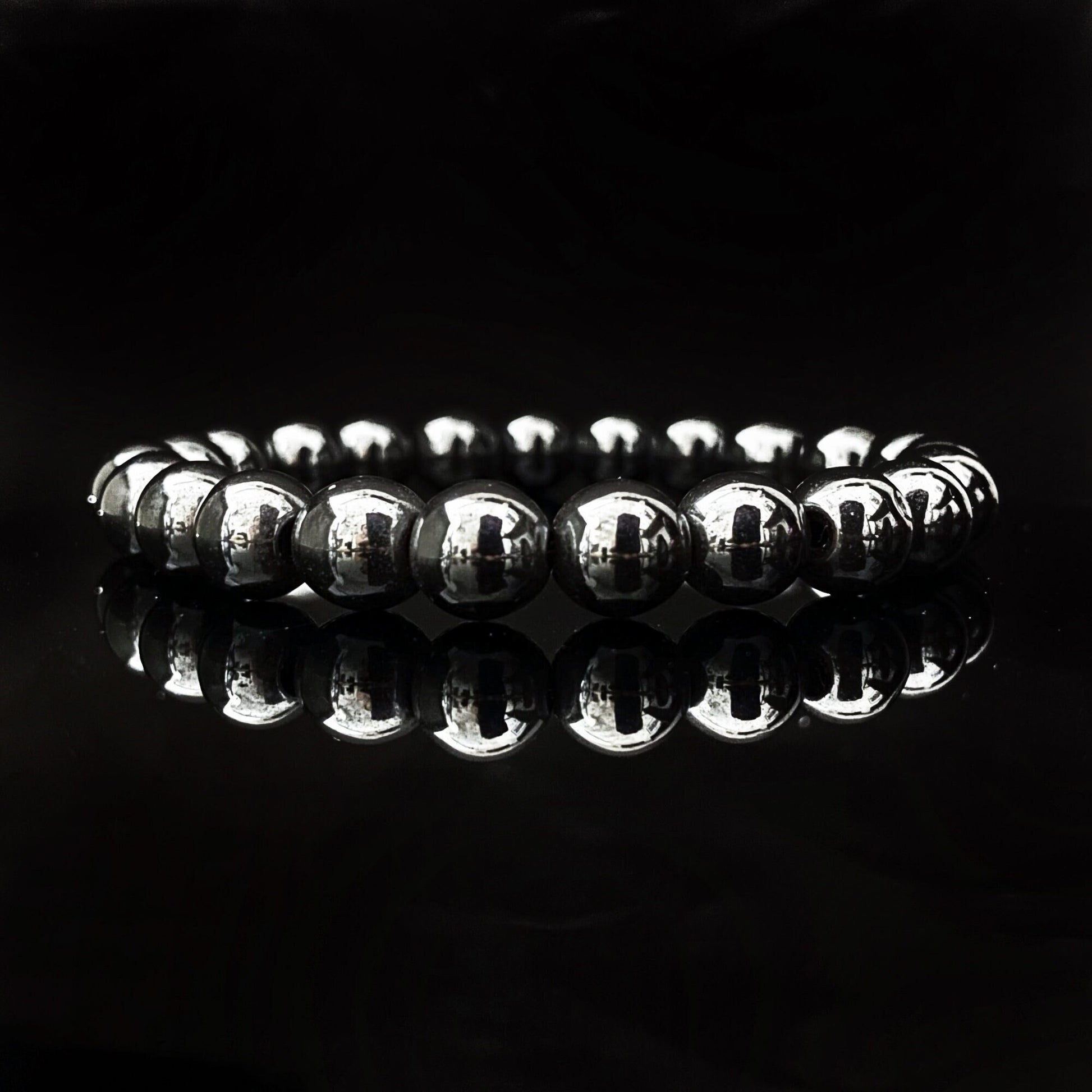 A reflective bracelet made of polished, spherical hematite beads arranged in a circle against a dark, glossy background. The lighting highlights the shiny surface of the 8mm Hematite Beaded Bracelet, creating a mirror-like reflection on the surface beneath and emphasizing its grounding properties.