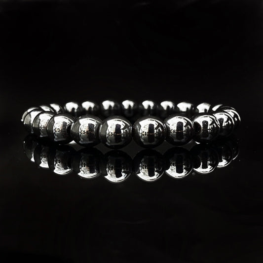 A reflective bracelet made of polished, spherical hematite beads arranged in a circle against a dark, glossy background. The lighting highlights the shiny surface of the 8mm Hematite Beaded Bracelet, creating a mirror-like reflection on the surface beneath and emphasizing its grounding properties.