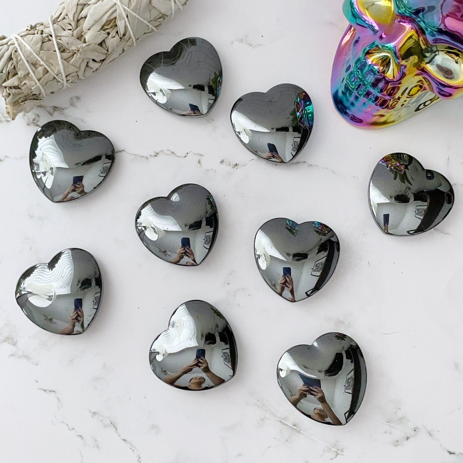 The 2" Hematite Heart Palm Stones are thoughtfully arranged on a marble surface alongside an iridescent skull and a sage bundle. The polished stones brilliantly reflect their surroundings, including the person taking the photo, crafting an image that exudes grounding presence and natural artistry.