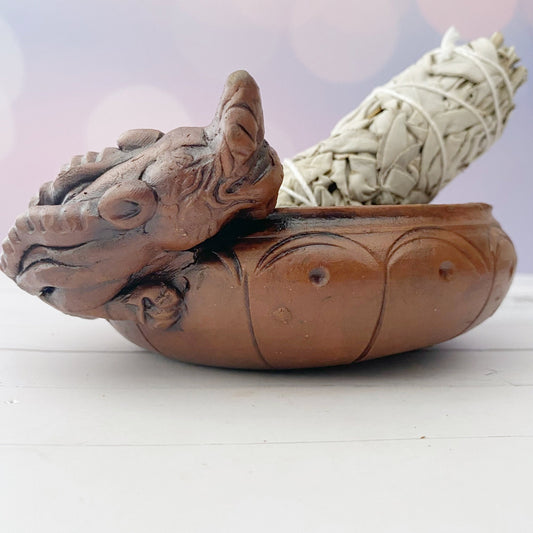 A decorative altar piece, this Terracotta Dragon Incense Burner features a striking dragon head design cradling a bundled sage stick. The background, slightly blurred with soft, warm lights, enhances the serene and calming atmosphere with an air of mythological creatures' mystique.