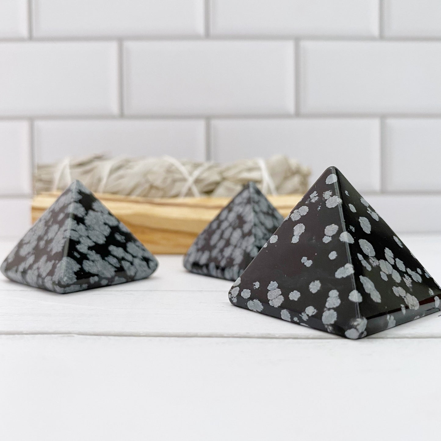 Three 1" Mini Snowflake Obsidian Pyramids are arranged on a white surface, accompanied by a wooden tray filled with bundles of dried herbs in the background. The pyramids feature black stone accented with light gray snowflake-like patterns, celebrated for their metaphysical properties. A tiled wall serves as the backdrop.