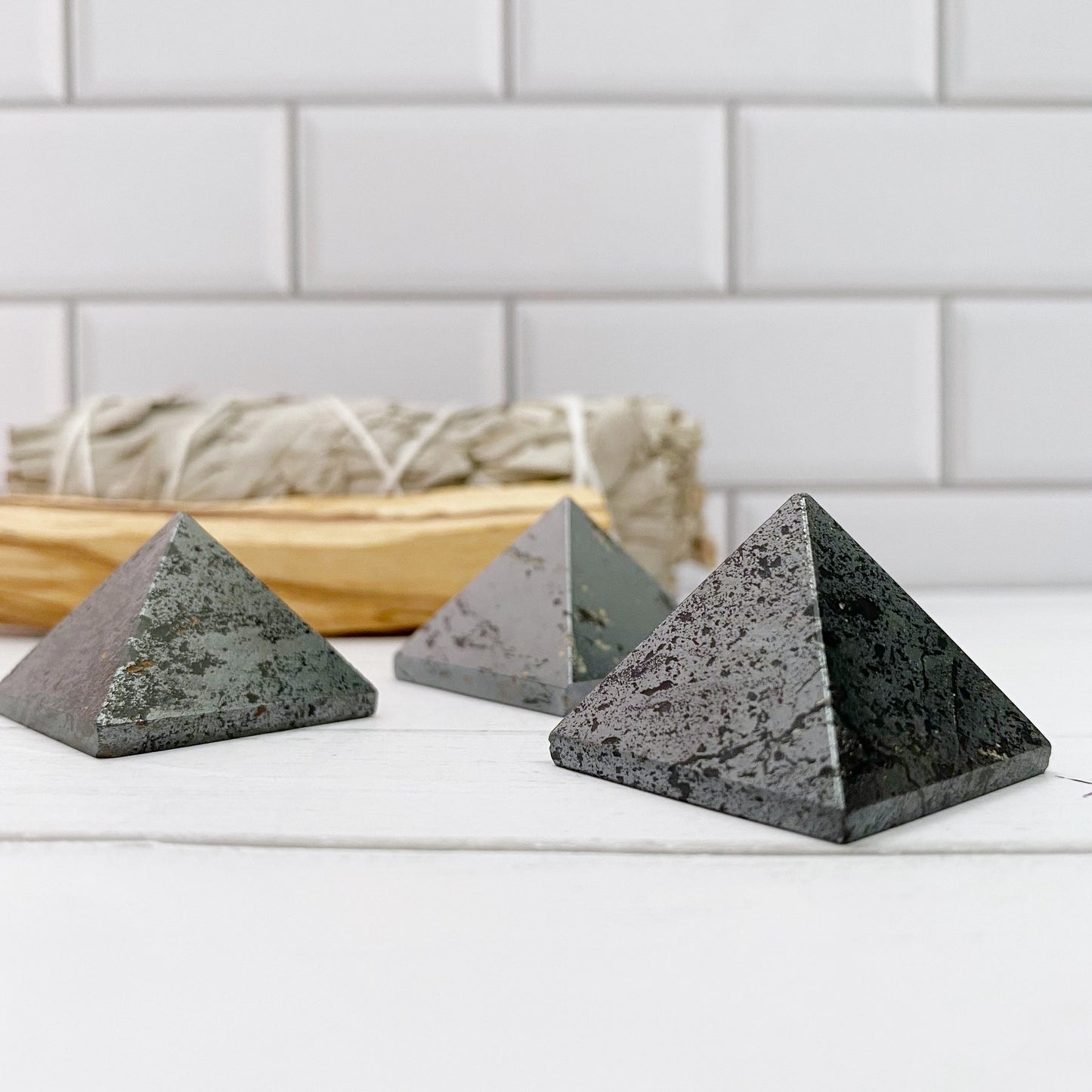 Three pyramid-shaped stones, including a 1" Mini Hematite Pyramid known for its grounding properties, are placed on a white surface with a bundle of sage in the background. The scene is set against white subway tiles, creating a clean and serene aesthetic perfect for meditation.