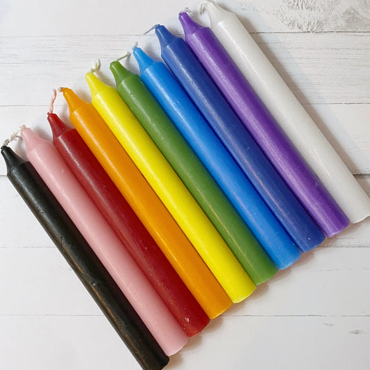 A collection of 12 colorful 5" Variety Pack Unscented Chime Candles (10 Pack) arranged horizontally on a white wooden surface. The candles display a variety of hues, including black, pink, red, orange, yellow, light green, dark green, light blue, dark blue, purple, and white—perfect for meditation or Chakra balancing.