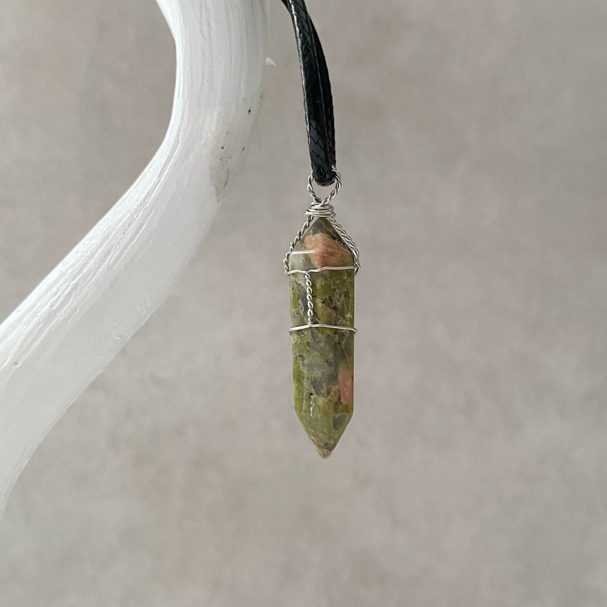 A 1.5" Double Terminated Unakite Point Pendant, showcasing a wire-wrapped greenish-brown crystal with hints of pink, is suspended from a black cord. Renowned for its metaphysical properties of spiritual healing, the pointed crystal pendant hangs from a white, textured branch against a blurred grey background.