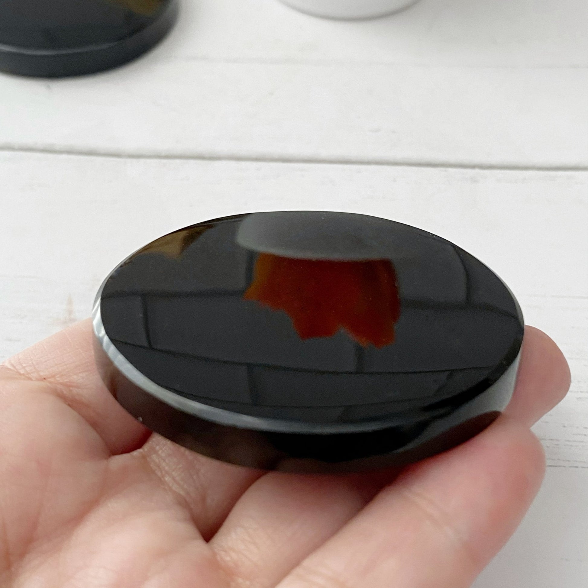 A 2" Black Obsidian Scrying Mirror with a reflection of a figurine of a mother and child. In the background, there's a small bundle of dried leaves tied together and an orange succulent plant in a white pot. The scene has a minimalist aesthetic perfect for divination practices.