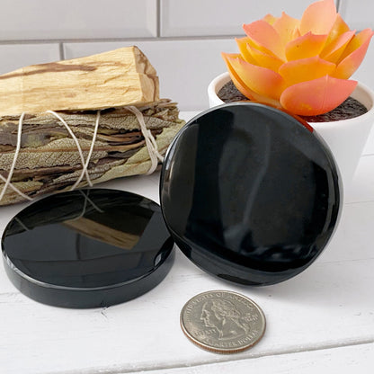 A 2" Black Obsidian Scrying Mirror with a reflection of a figurine of a mother and child. In the background, there's a small bundle of dried leaves tied together and an orange succulent plant in a white pot. The scene has a minimalist aesthetic perfect for divination practices.