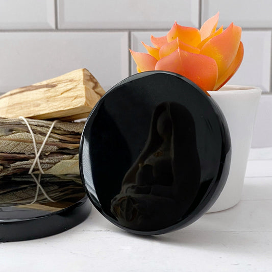 A 2" Black Obsidian Scrying Mirror with a reflection of a figurine of a mother and child. In the background, there's a small bundle of dried leaves tied together and an orange succulent plant in a white pot. The scene has a minimalist aesthetic perfect for divination practices.