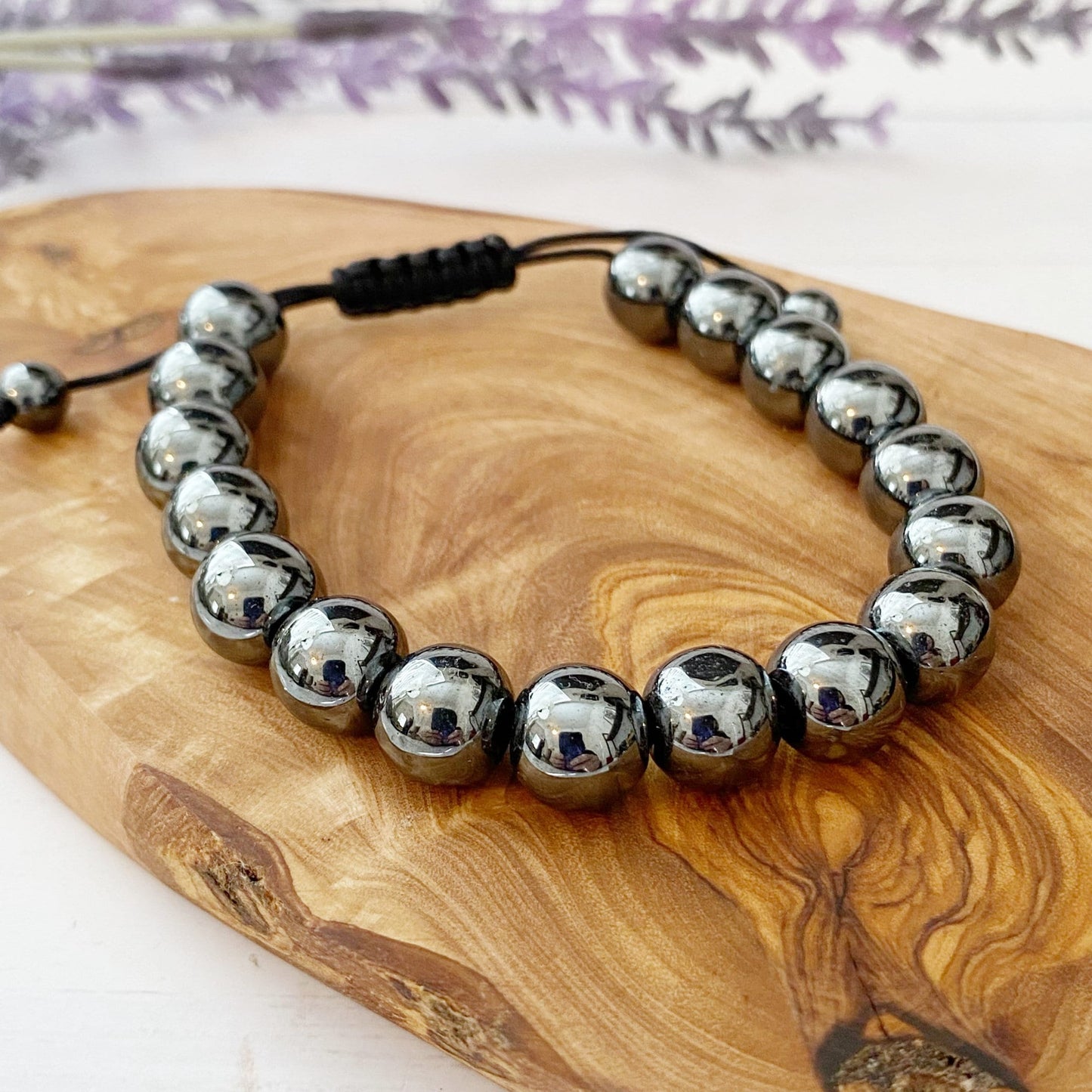 A 10mm Hematite Beaded Bracelet made of polished metallic beads, threaded onto an adjustable black cord. The shiny beads reflect light, creating a sleek and modern appearance with universal appeal. The bracelet is coiled into a circular shape, with the black cord ends visible at the top.