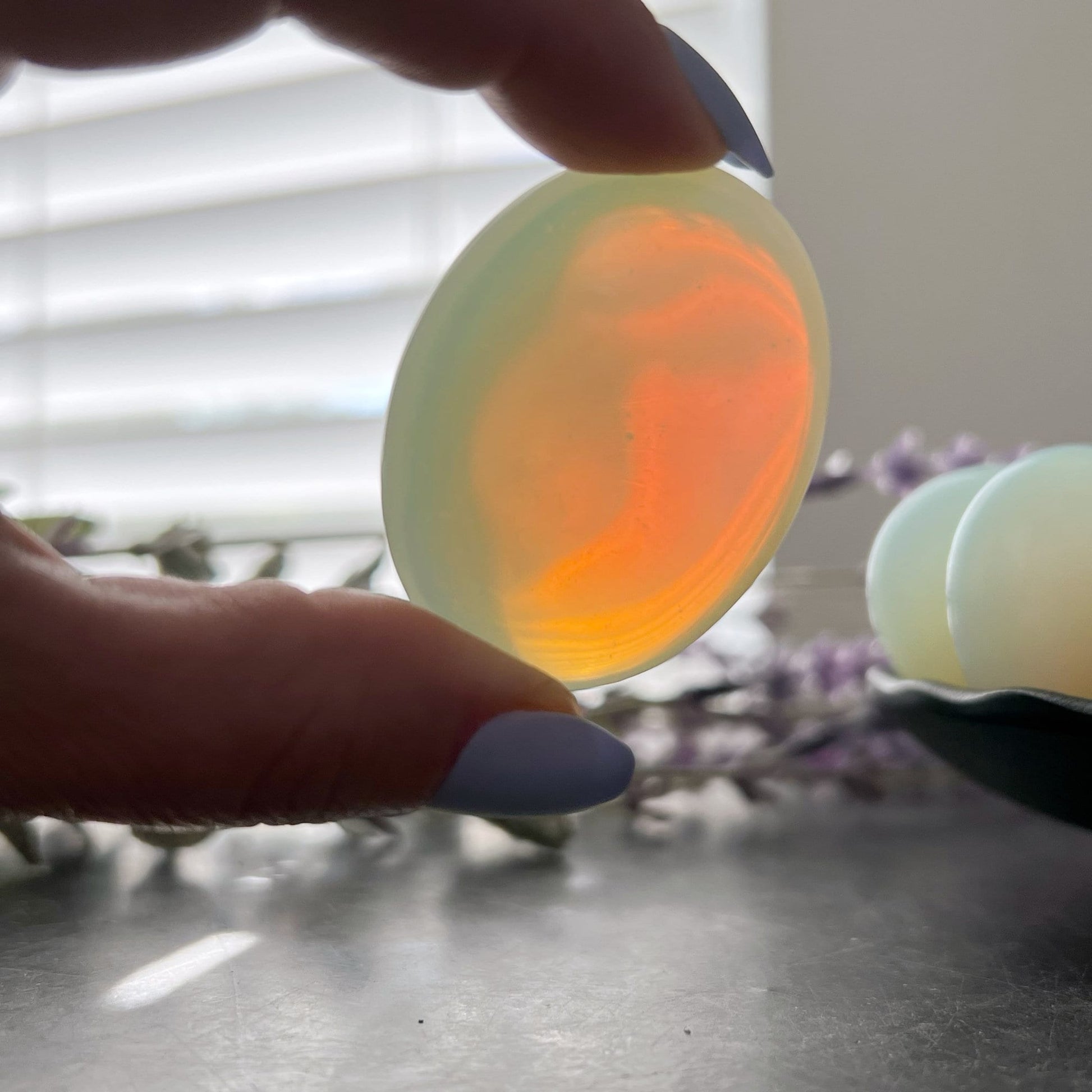 A black dish is arranged with smooth, oval-shaped 1.5" Opalite Palm Stones, radiating like petals around a central pair. Known for their healing properties, these milky white gemstones sit gracefully beside lavender sprigs on a gray surface.