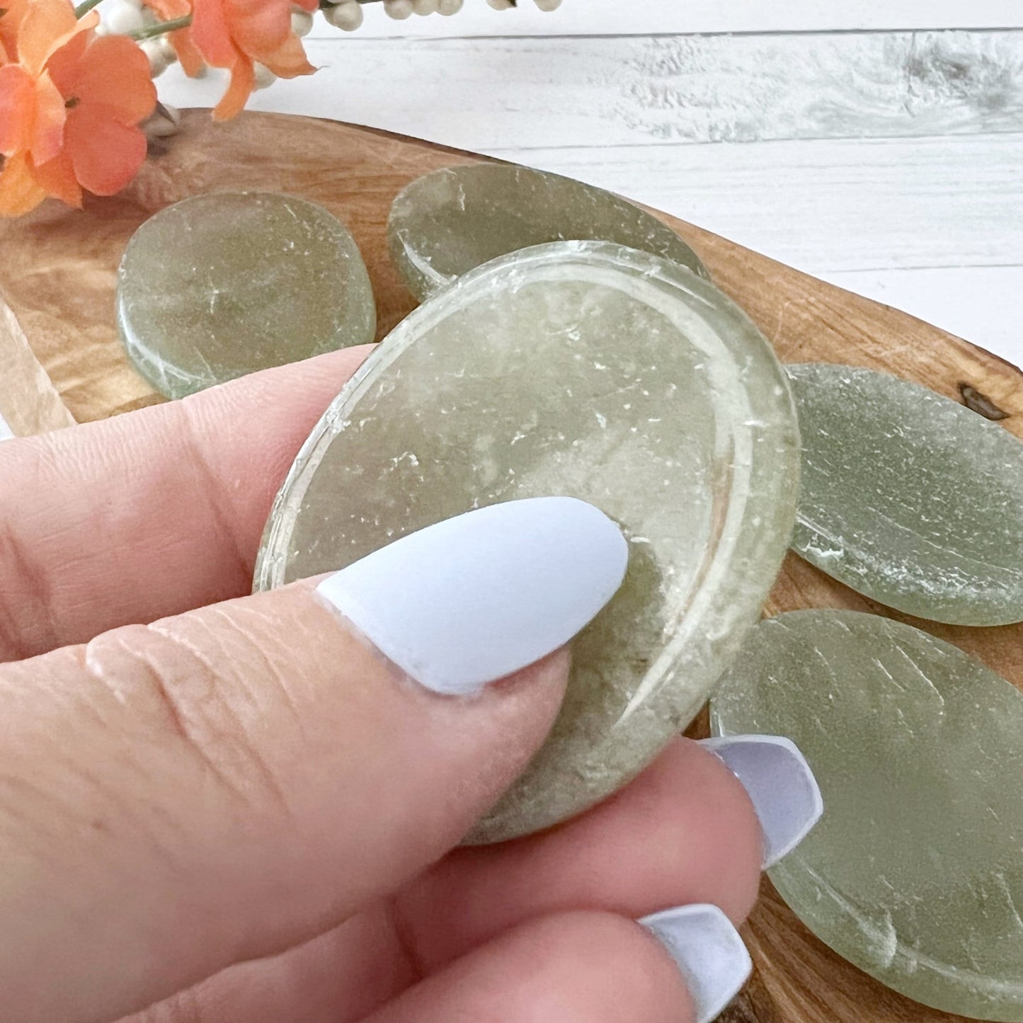 Four polished Green Aventurine Worry Stones are displayed on a wooden tray. One additional crystal rests off the tray on a white surface. A green leaf and an orange flower adorn the top left corner of the image, offering both natural beauty and stress relief for your senses.