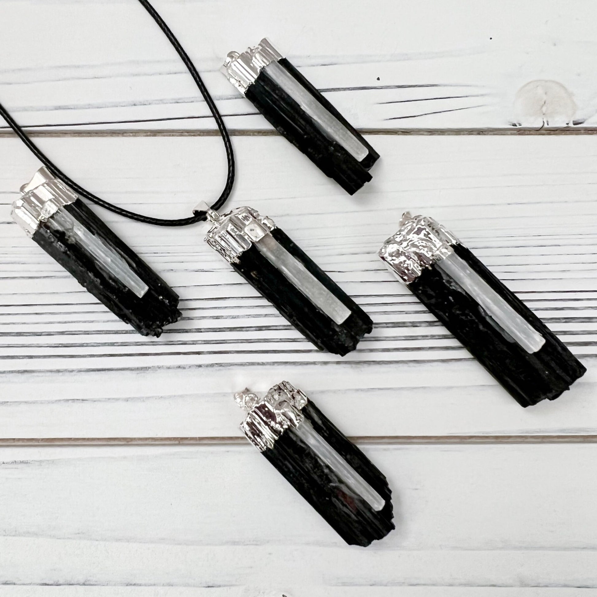 A Raw Black Tourmaline & Selenite Pendant hangs from a thin black cord. The rectangular stone has a rough, textured surface with silver accents at the top, emanating protective energy. The background is a simple, neutral grey.