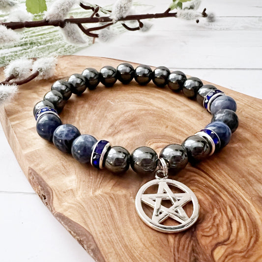 A beaded bracelet featuring blue sodalite and black hematite beads with silver accents and a pentacle charm rests on a wooden surface. The background includes a soft green and white floral element, adding a natural touch to the 8mm Hematite & Sodalite Beaded Bracelet with Pentacle Charm composition.