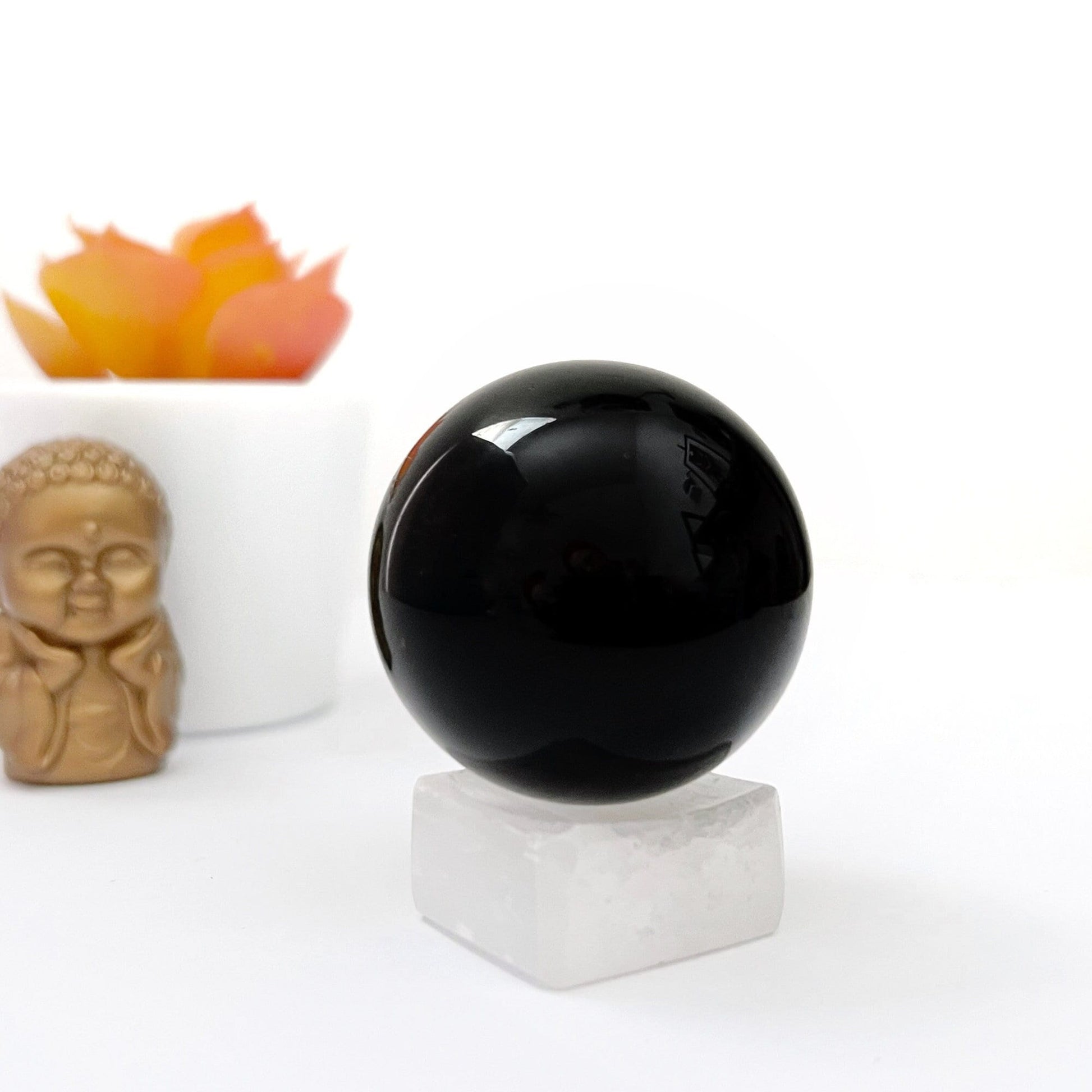 A 40mm Obsidian Crystal Ball rests on a white cubic stand with a small statue of a meditating figure and a potted succulent in the background. The scene, set against a white backdrop, evokes an atmosphere perfect for meditation and divination.