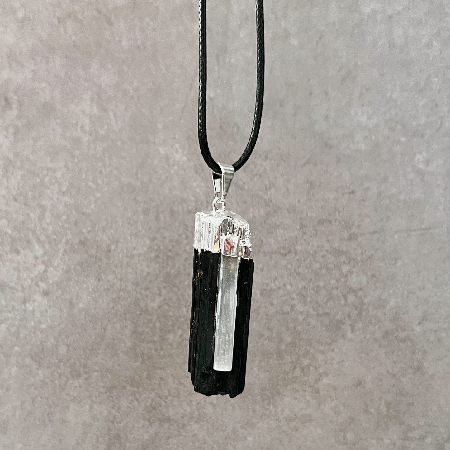 A Raw Black Tourmaline & Selenite Pendant hangs from a thin black cord. The rectangular stone has a rough, textured surface with silver accents at the top, emanating protective energy. The background is a simple, neutral grey.