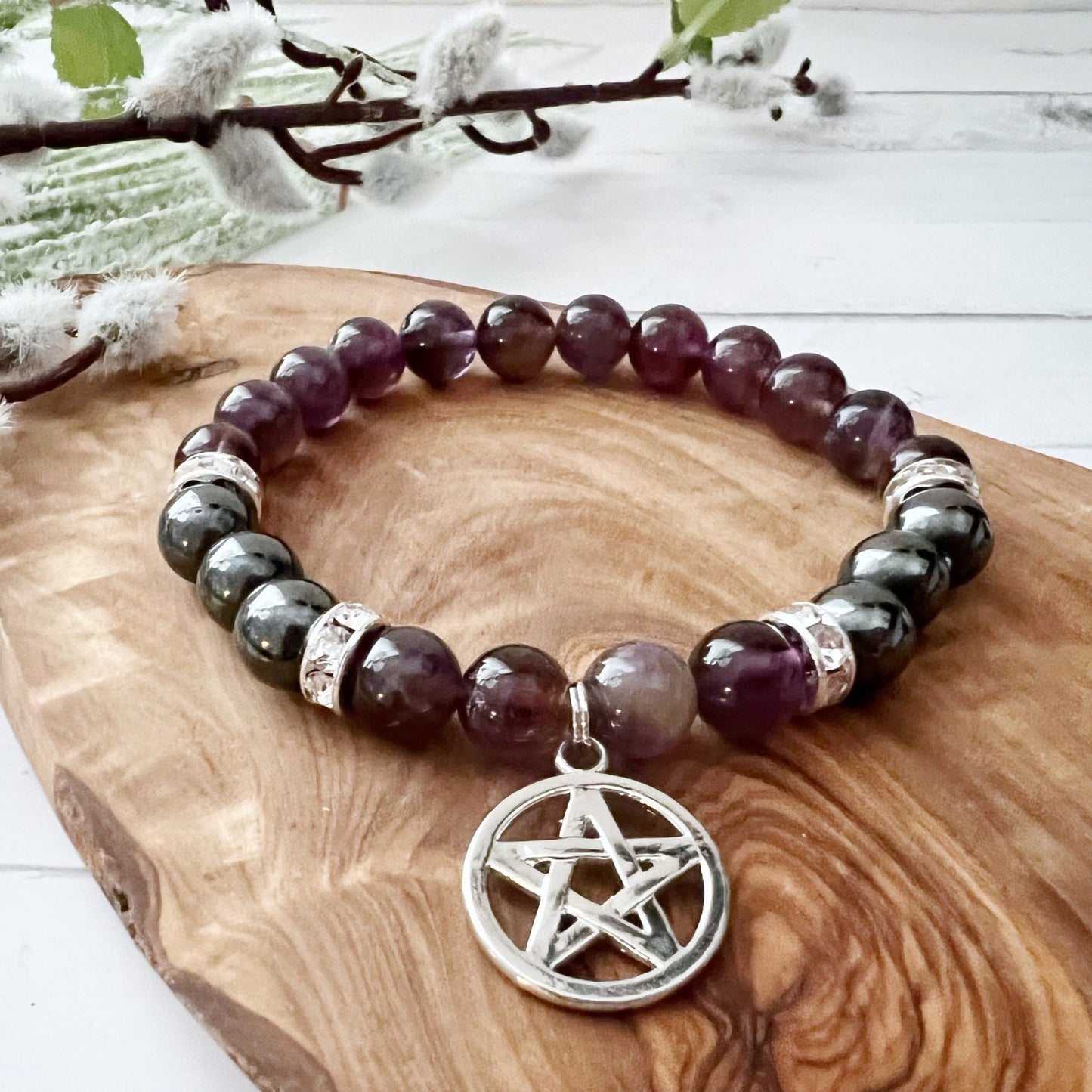 This image features an 8mm Hematite & Amethyst Beaded Bracelet with Pentacle Charm with alternating purple and dark gray beads, adorned with silver accents. The bracelet has a silver pentacle charm attached. It is placed on a wooden surface next to a branch with green and white-budded twigs.