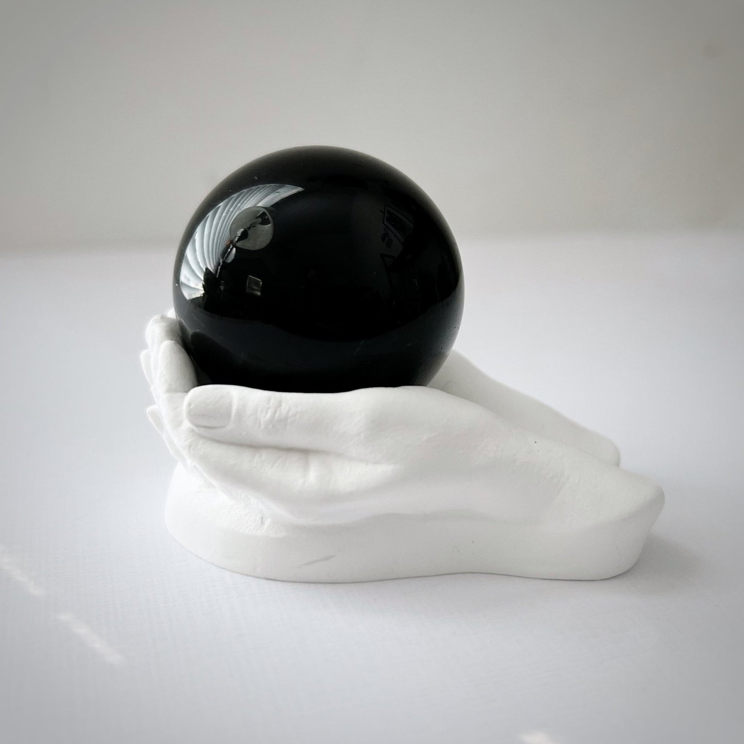 A 40mm Obsidian Crystal Ball rests on a white cubic stand with a small statue of a meditating figure and a potted succulent in the background. The scene, set against a white backdrop, evokes an atmosphere perfect for meditation and divination.