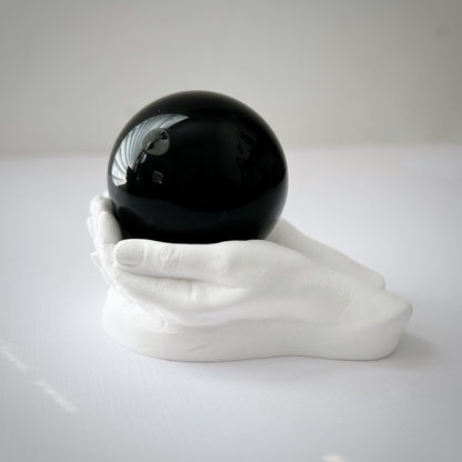 A 40mm Obsidian Crystal Ball rests on a white cubic stand with a small statue of a meditating figure and a potted succulent in the background. The scene, set against a white backdrop, evokes an atmosphere perfect for meditation and divination.