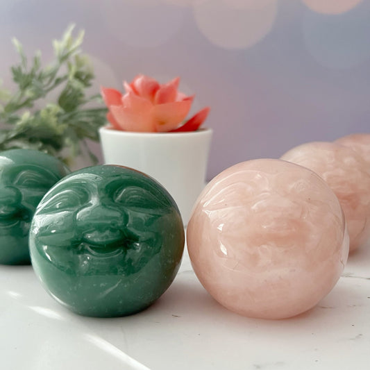 A close-up image of two 2" Man on the Moon / Buddha Carved Moons with smiling faces, one Green Aventurine and one Rose Quartz, on a white surface. In the background, there's a small green plant and a pink succulent in a white pot, with a blurred light purple backdrop.