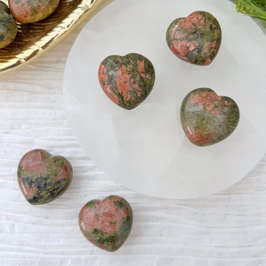 Five 1" Mini Unakite Pocket Hearts, displaying a mix of pink and green hues, are arranged on a round white plate placed on a textured white surface. A golden tray with similar stones, noted for their ability to promote emotional balance and grounding energy, is partially visible on the left side of the image.