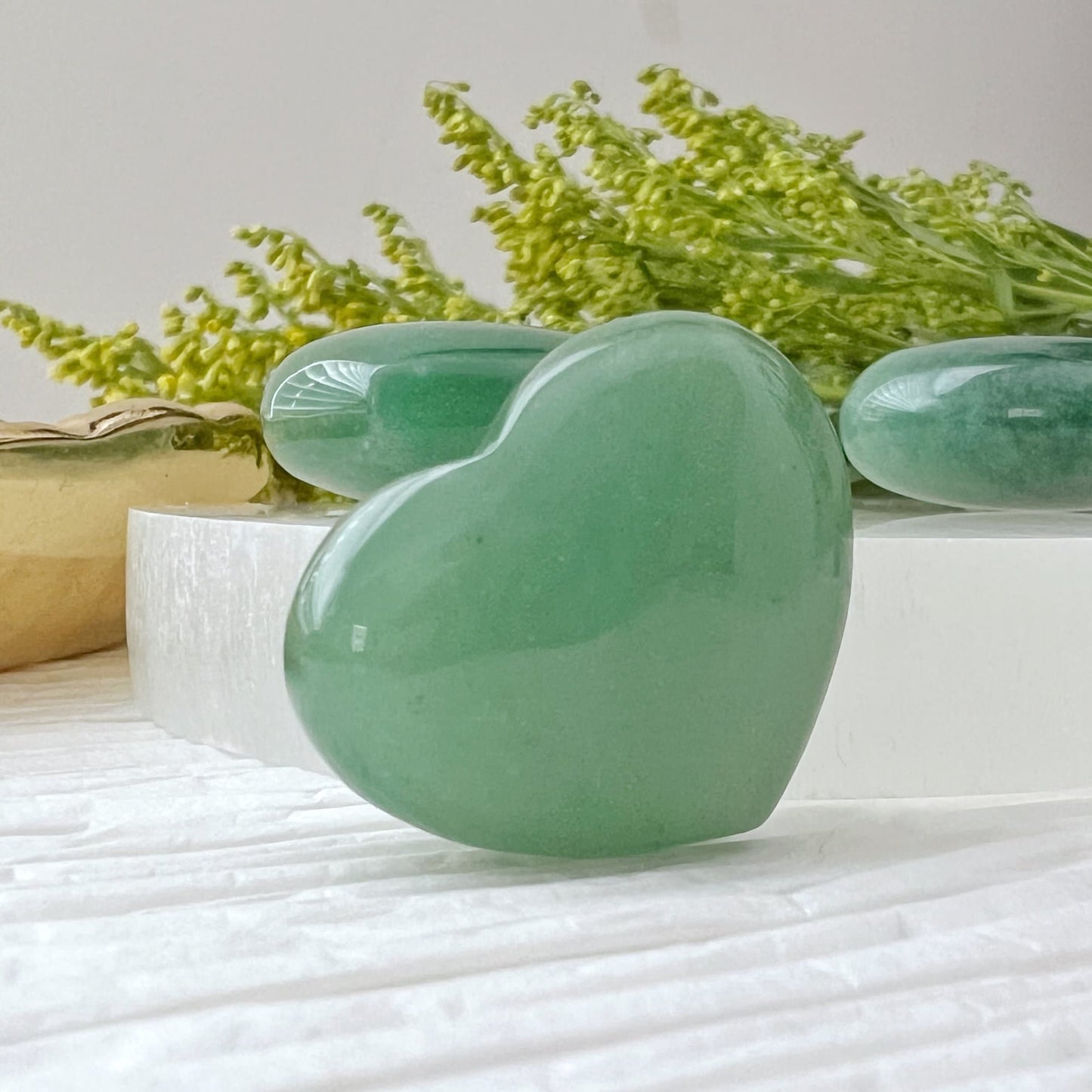 A collection of green heart-shaped 1" Mini Green Aventurine Pocket Hearts is displayed on a white surface. Some stones are placed on a circular white tray, while others rest on a gold-colored leaf-shaped tray. Green foliage partially frames the upper right corner of the image, manifesting prosperity and abundance.