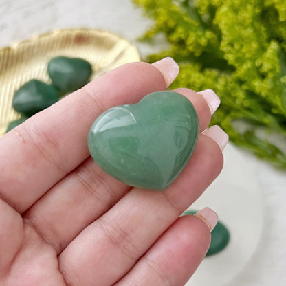 A collection of green heart-shaped 1" Mini Green Aventurine Pocket Hearts is displayed on a white surface. Some stones are placed on a circular white tray, while others rest on a gold-colored leaf-shaped tray. Green foliage partially frames the upper right corner of the image, manifesting prosperity and abundance.