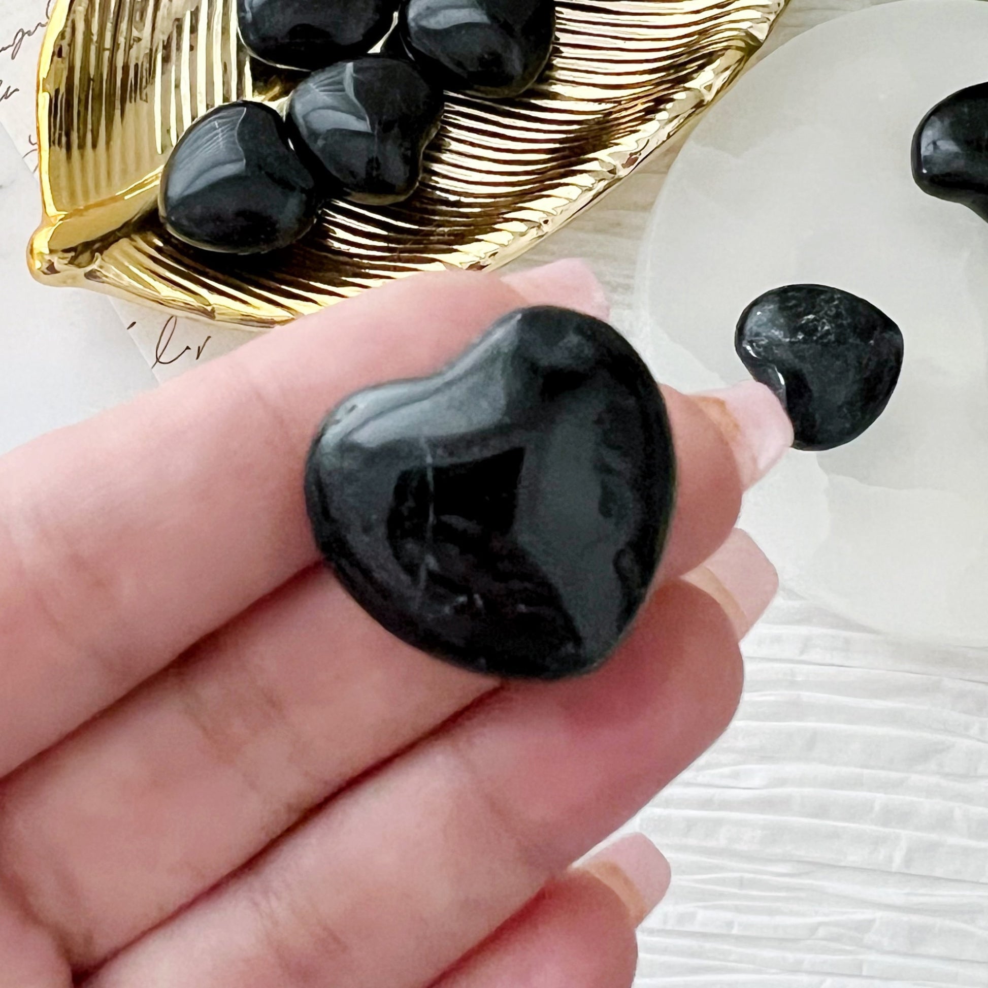 A collection of small, polished 1" Mini Black Tourmaline Pocket Heart stones are arranged on a light circular surface, with more of the same stones in a golden dish next to it. Green foliage and textured paper are visible in the background, offering negative energy protection.