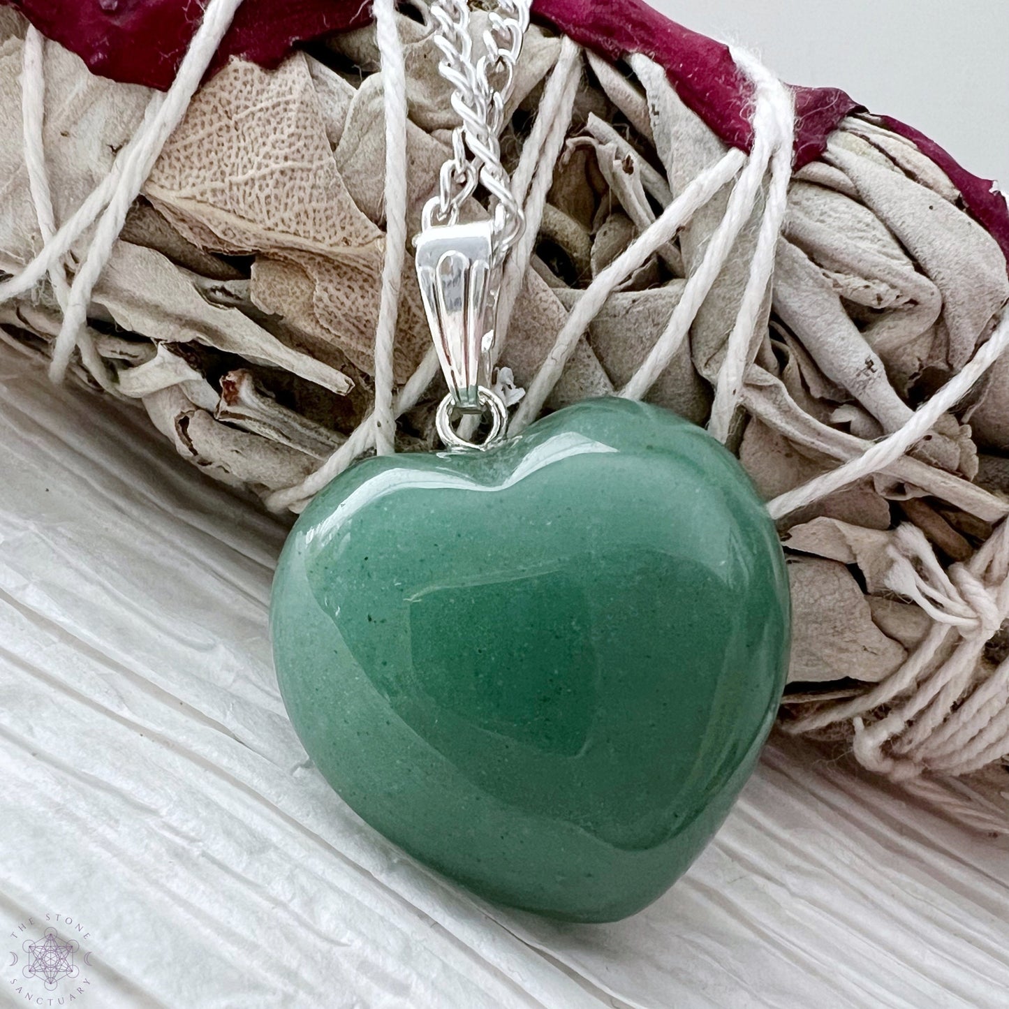 A silver necklace with a 1" Green Aventurine Heart Pendant is displayed on a wooden slab alongside three matching green heart-shaped pendants. To the left, there is a blush pink flower. The background is a textured white surface. This healing stone accessory exudes both elegance and tranquility.