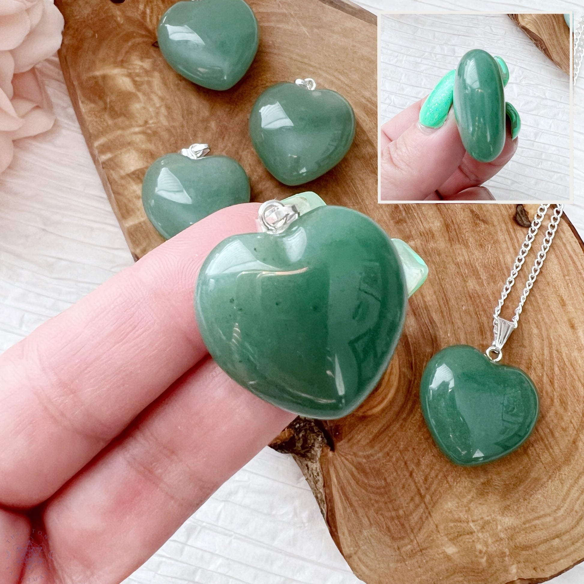 A silver necklace with a 1" Green Aventurine Heart Pendant is displayed on a wooden slab alongside three matching green heart-shaped pendants. To the left, there is a blush pink flower. The background is a textured white surface. This healing stone accessory exudes both elegance and tranquility.