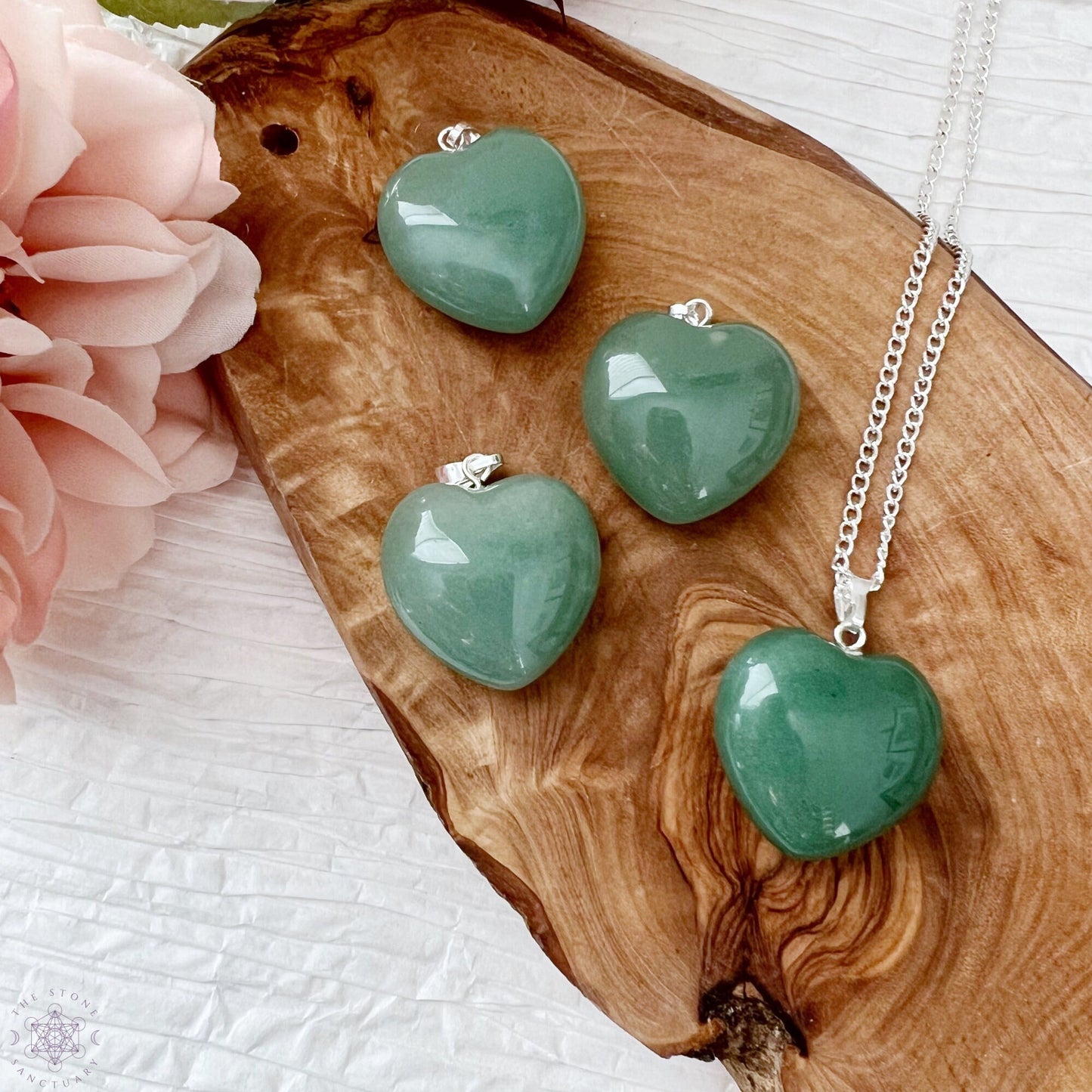 A silver necklace with a 1" Green Aventurine Heart Pendant is displayed on a wooden slab alongside three matching green heart-shaped pendants. To the left, there is a blush pink flower. The background is a textured white surface. This healing stone accessory exudes both elegance and tranquility.