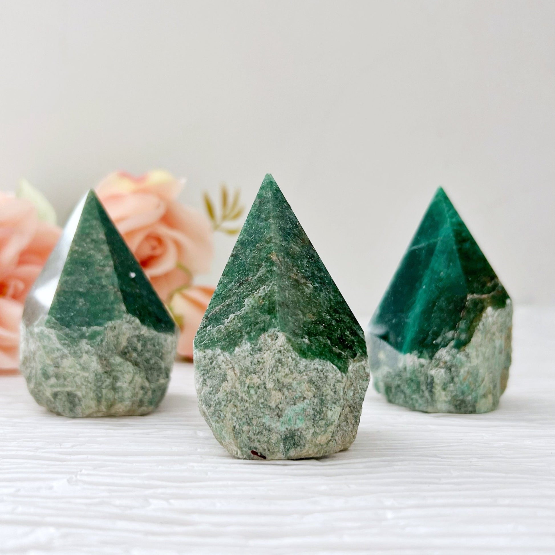 Three 2-3" Green Aventurine Top Polished Points, perfect for heart-centered healing, stand upright on a textured white surface. The crystals have a dark green top and a lighter, rougher base. In the background, there are some blurred peach-colored roses.